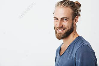Happy young bearded guy with fashionable hairstyle and beard looking at camera, brightfully smiling with teeth, being