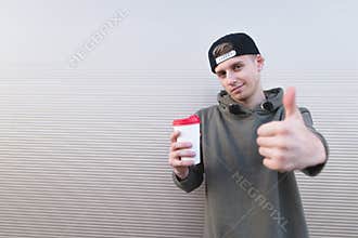 A stylish young man with a cup of coffee in his hands shows a thumbs up on a light background.