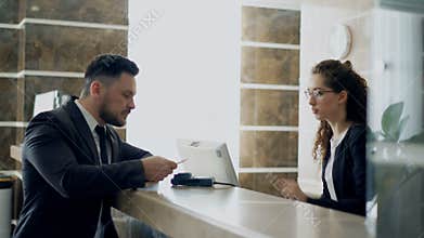 Businessman guest talking with receptionist girl standing at desk in hotel reception and paying bill with credit card