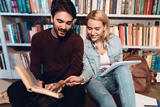 White guy and girl near bookshelf in library. Students are reading books.