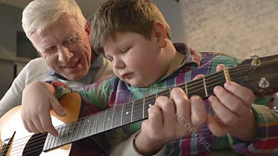Grandfather is teaching his grandson how to play the guitar. An elderly man is teaching a young fat child to play guitar