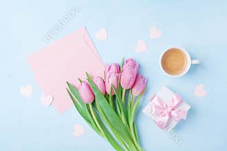Coffee cup, spring tulip flowers, gift box and pink paper card on blue pastel table top view. Greeting for Womans or Mothers Day.