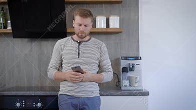 Handsome man putting cup in coffee-maker and switching it on his kitchen morning, then typing messages in his smartphone