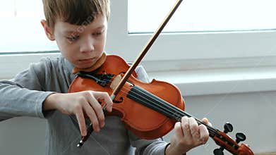 Play the violin. Seven years old boy playing the violin near a window. Front view.