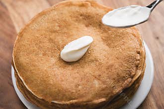 Adiing sour cream to stack of fresh hot crepes or blinis on wood table