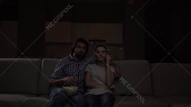 Man and woman sit together and watch TV. Guy embracing girl. They eat popcorn. Movie is very excited. They are