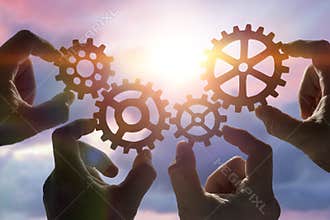 Four hands collect a puzzle of gears, against the background of the sky at sunset.