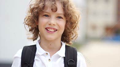 Preteen curly blue-eyed boy laughing and looking at camera. The schoolboy is dressed in a white shirt and carries a
