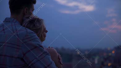 Young guy gently hugging his girlfriend by shoulders warming her at sunset