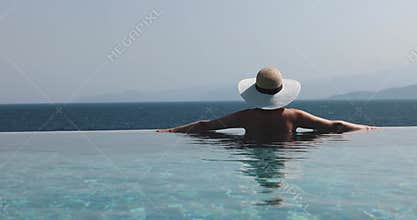 Woman with straw hat relaxing in infinity swimming pool and enjoying sea view at luxury resort