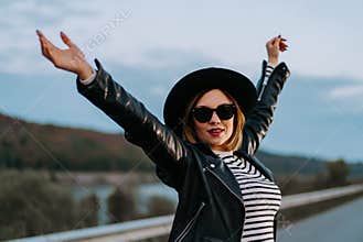 Portrait of young carefree woman standing on empty night street with open arms. Girl in striped top and leather jacket