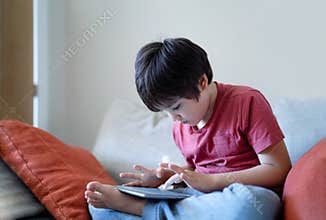School kid sitting on sofa watching cartoon on tablet, Cute young boy playing game online on digital pad, Cinematic indoor