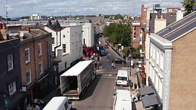 Fly over streets in urban neighbourhood. Pedestrians walking along parked vans and cars. Truck unloading. London, UK