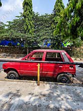 Stock photo of wreaked red color car, broken and rusty car parked near pathway in the Bangalore city . Green trees on background,