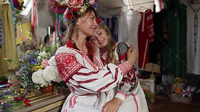 Happy mother and daughter in national Ukrainian costumes sitting indoors hugging looking away. Portrait of beautiful