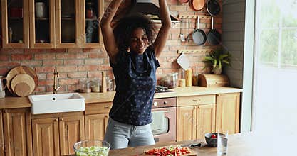 Joyful active millennial Black housewife dance at kitchen enjoy cooking