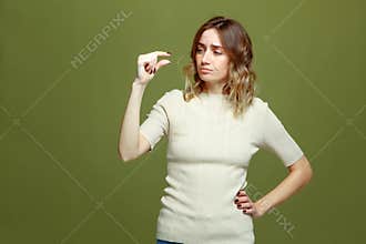 Small amount gesture. Sad dissapointed young woman showing little size by fingers on green studio background