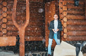 Man staying in the country house porch and enjoying snowfall. Winter countryside holidays concept image