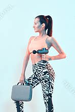 Athletic young female in sportswear posing with handheld massage gun for post-workout recovery in neon studio light