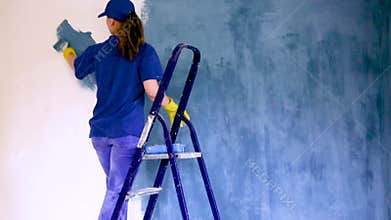 A young woman in a blue T-shirt in yellow rubber gloves and cap is painting a wall in an apartment, there is a staircase nearby.
