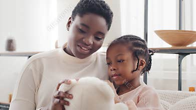 Afro american mother with little daughter girl sitting on sofa at home talking holding teddy bear. Single parent ethnic