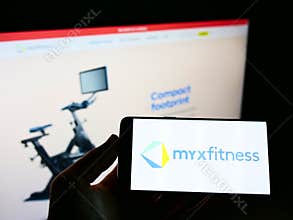 Person holding cellphone with logo of American fitness streaming platform MYXfitness on screen in front of company webpage.