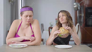Jealous fat woman looking at slim Caucasian friend eating sandwich. Front view of slender and obese women sitting at