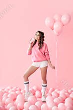 Full length portrait of trendy young woman drinking milk shake over pink balloons background