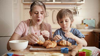 Smiling and laughing mother with little son eating freshly baked croissants on messy kitchen at home. Children cooking
