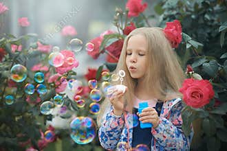 Girl blowing rainbow color soap bubbles in the bush of roses