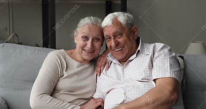 Happy older spouses sit on couch hug look at camera