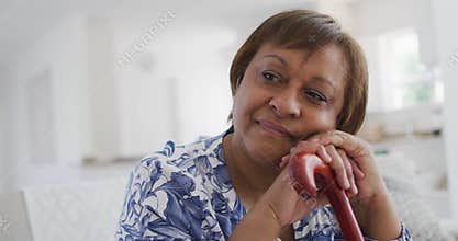 Happy african american senior woman sitting leaning on walking stick, looking away smiling