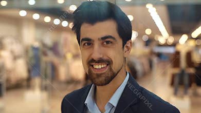 Close-up face of handsome bearded young man looking at camera and smiling standing in modern light shopping mall.
