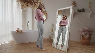 Displeased lovely adult woman trying to button tight denim in front of mirror after gaining weight