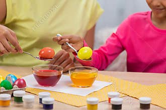 Mom and daughter with funny bunny ears dips eggs in food coloring using yellow and red paint. African American woman and