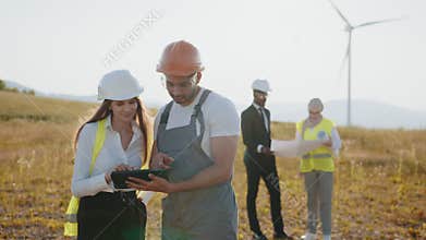 Four colleagues using blueprints and tablet outdoors. Multiracial colleagues controlling work of windmill farm. Eco for
