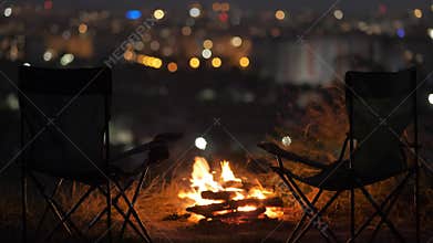 Two empty camping chairs near burning yellow camp fire at night.