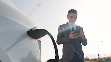 Caucasian businessman using smart phone and waiting power supply connect to electric vehicles for charging the battery