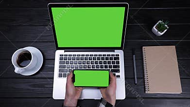 The girl works using several devices, laptop, smartphone. Top view of a laptop with a green screen and a phone with a