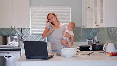Mother multi-tasking, holding baby infant and using computer laptop at home. Candid authentic and real life mom working