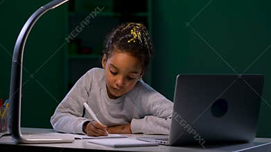 A girl using a laptop listens to a video tutorial and takes notes in her notebook. A child sits at a table in a dark