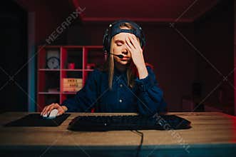 Upset girl gamer sits at night at the computer and shows a gesture of facepalm, lowered his eyes down from the shame of losing the