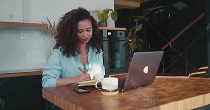 Happy young attractive African American entrepreneur business woman taking notes, using laptop at home kitchen table.