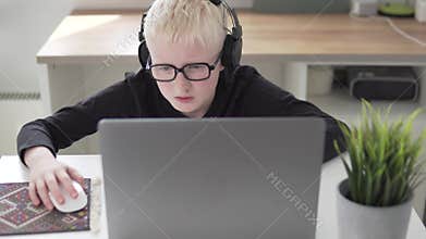 A boy with glasses and headphones plays in uses a laptop at home at the table