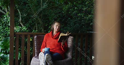 Senior caucasian woman relaxing in garden, sitting with feet up, drinking coffee and reading book