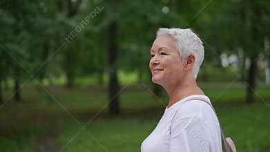 Closeup tracking shot of happy elderly 65s female walking through natural park with green foliage. Smiling elderly