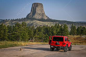 Red Jeep parked near Devils Tower National Monument with a small dog sitting on the ground