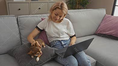 A young blonde woman is sitting in a living room, working on a laptop and petting her chihuahua on the couch