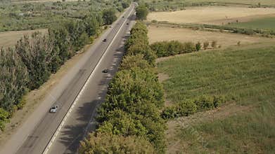 White, gray and black cars drive along a four-lane paved highway past meadows, fields and green trees.