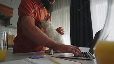 A young bearded man in a good mood stands in the kitchen, uses a laptop to searching and typing information, writes